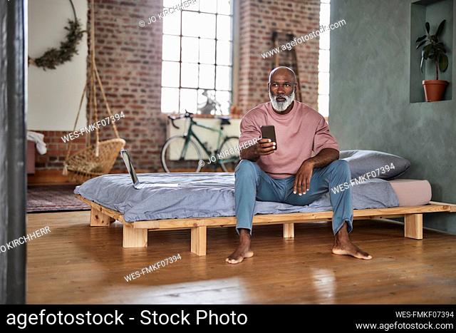 Bald man with smart phone sitting by laptop on bed at home