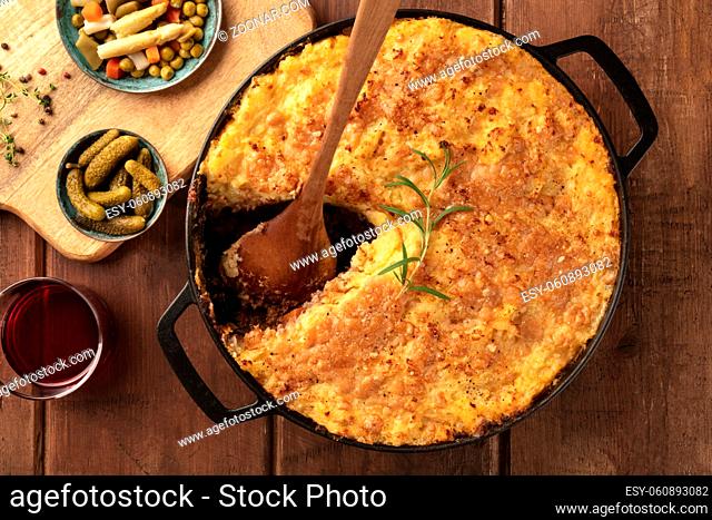 Homemade Shepherd's pie in a cooking pan with pickles, herbs, and wine, with a missing slice, overhead shot on a dark rustic wooden background