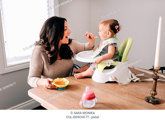Mother feeding baby daughter in child seat on kitchen table