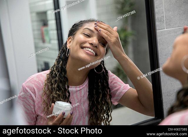 Happy young woman applying moisturizer in bathroom mirror