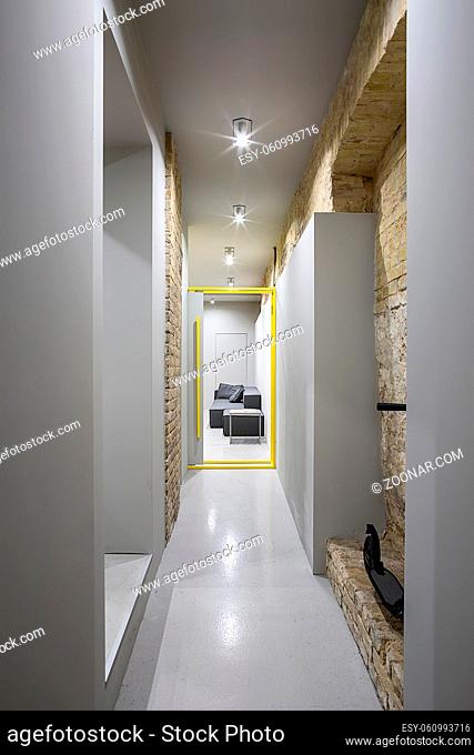 Loft style corridor with a glowing lamps and brick and gray walls. There is a opened gray-yellow door to the room with a dark sofa and a small wooden table