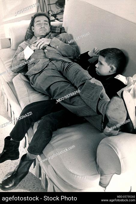 Italian actor Ugo Tognazzi sleeping on the couch with his legs raised and leaned over the legs of his son Ricky. 1960s