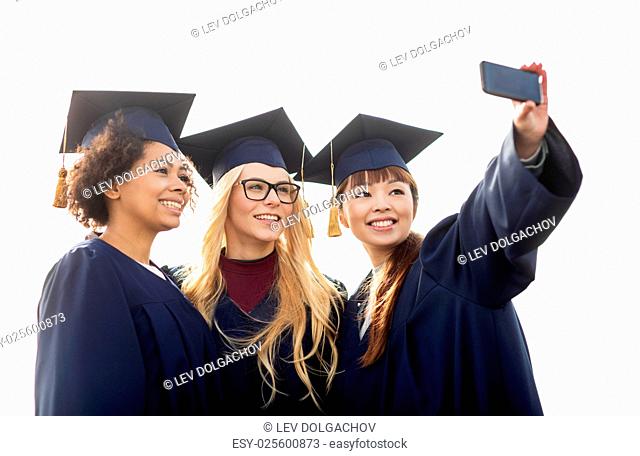 education, graduation, technology and people concept - group of happy international students in mortar boards and bachelor gowns taking selfie by smartphone...