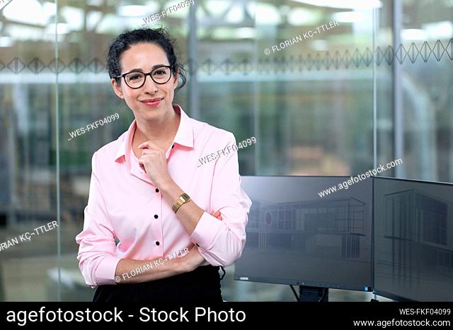 Smiling businesswoman standing by computers against glass wall at office
