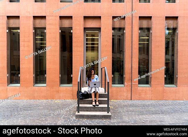 Businesswoman using laptop while sitting on steps at entrance of office building