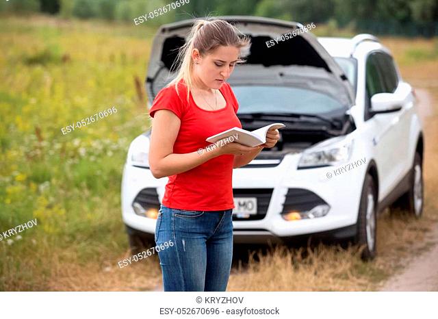 Young stressed woman standing at broken car and reading owner manual