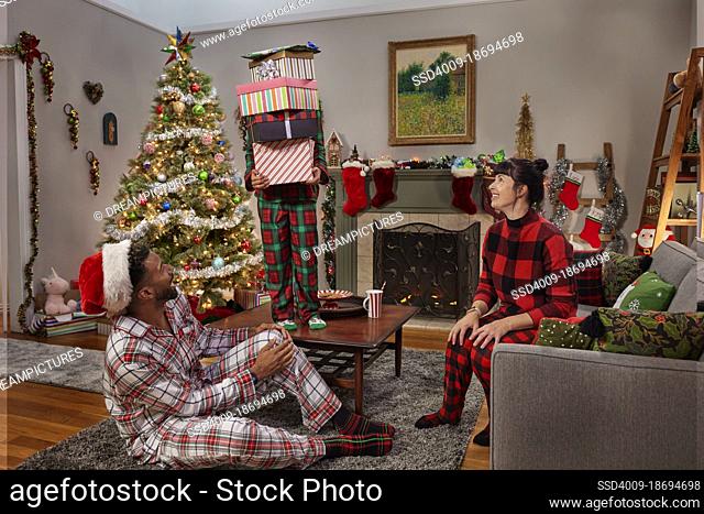 Girl holding tall stack of Christmas presents standing of coffee table as her parents look on laughing. Diverse Family