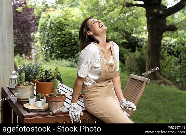 Cheerful young woman laughing while sitting on table in yard during curfew