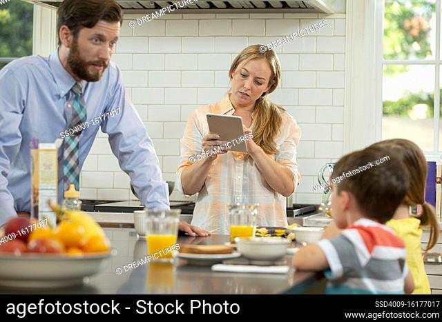 Mom with family in kitchen, struggling to get the kids breakfast and ready to start the day. Checking tablet to connect with doctor
