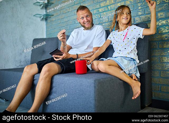Father and daughter testing talking smart wireless speaker, controlling home devices with a voice commands, sitting on sofa at home, Concept of smart home