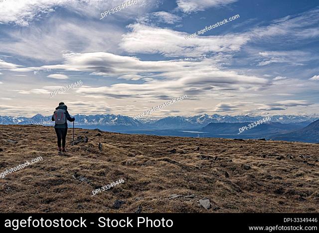 View taken from behind of a woman hiking with trekking poles on the rugged vegetation and rocks of the tundra in front of a silhouetted mountain range with a...