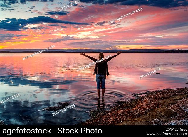 Female with arms outstretched enjoys the sunset turning the sky shades of red and mirrored reflecting colours in the water