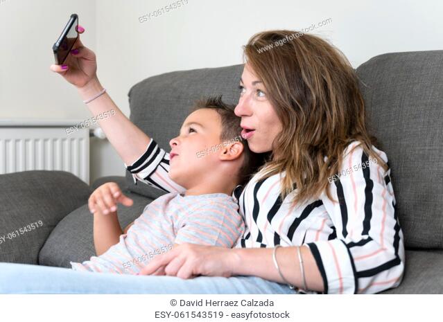 Cheerful mother holding smart phone making selfie with her little son showing victory sign. mom and kid boy sit on couch using gadget having fun taking...
