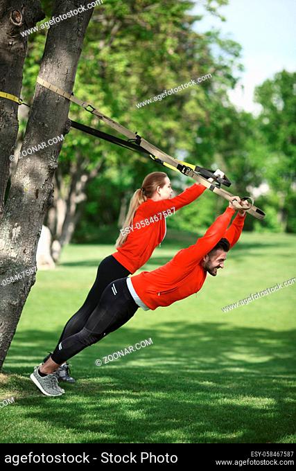 Couple training with suspension trainer sling in green park near spring or summer tree. Man and woman in red jackets preparing for sports competitions