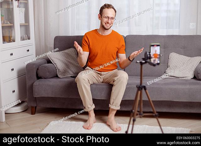 Hipster man blogger recording vlog on mobile phone sit on couch in living room and explain and gesturing with hands