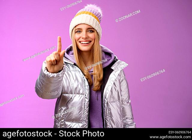 Excited cheerful fair-haired european girl in winter hat silver shiny jacket show number one index fingers give suggestion advice smiling broadly talking...
