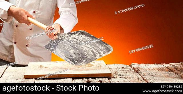 Chef holding a floured empty metal pizza paddle over an old rustic or vintage wooden kitchen table for food placement in a panorama banner with copy space