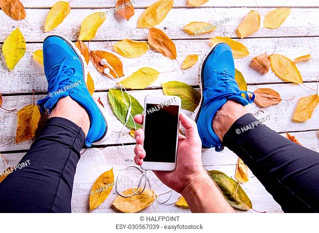 Legs of unrecognizable runner in blue sports shoes. Smart phone and earphones. Colorful autumn leaves. Studio shot on white wooden background