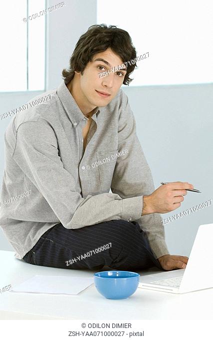 Man sitting with laptop, holding pen, looking at camera