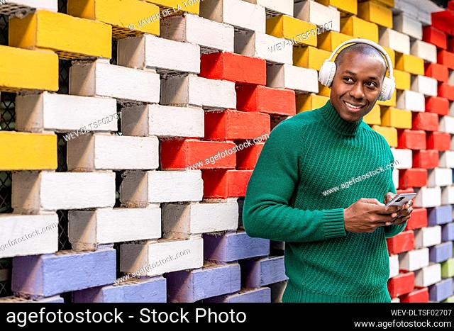 Smiling bald man with smart phone listening music through wireless headphones in front of multi colored wall