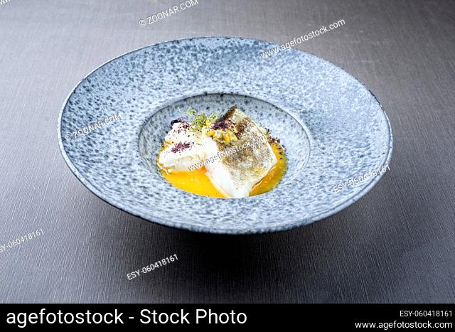 Modern style traditional sauteed skrei cod fish filet with skin with creme fraiche quenelles and algae in passion fruit sauce as close-up in ceramic design...