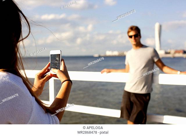 Young woman photographing boyfriend by pier over sea