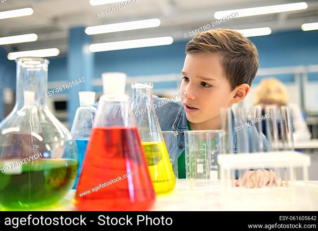 Interest. Surprised boy carefully examining flasks with colored liquids standing on table in chemistry class at school