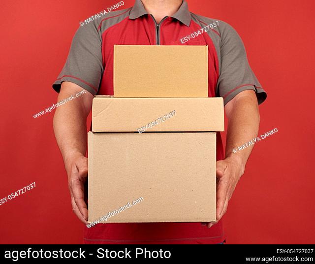 adult man in a T-shirt holds a stack of large cardboard brown boxes on a red background, concept of delivery of goods, moving