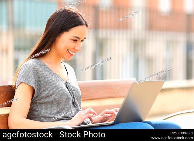 Happy woman smiling using laptop sitting on bench in the street