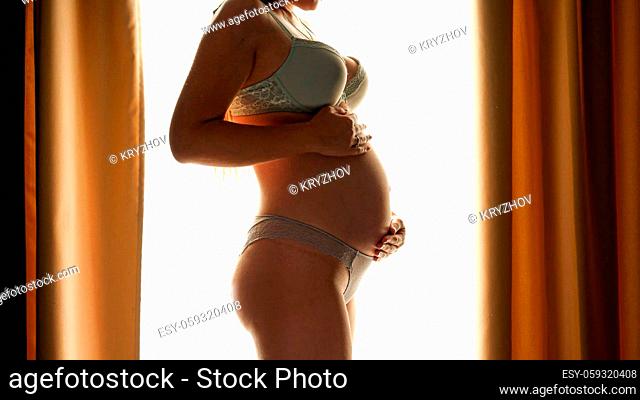 Silhouette of pregnant woman in lingerie stroking her big belly and touching tummy against big window in bedroom. Concept of happy pregnancy and baby...