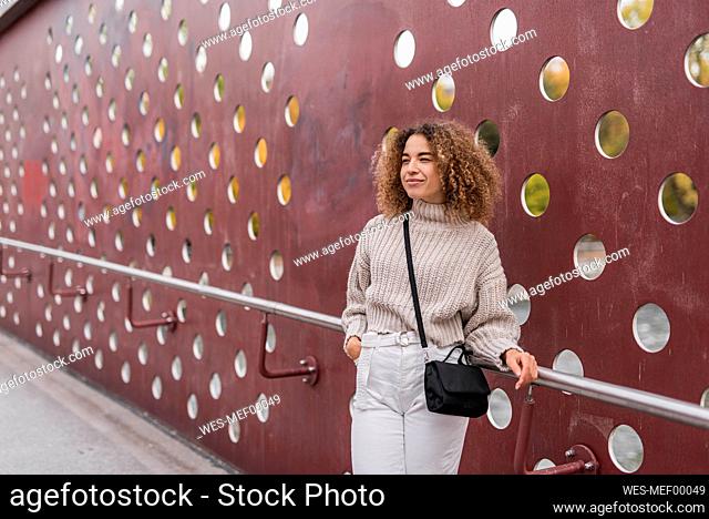 Beautiful young blond woman standing with hand in pocket while leaning on railing against metallic wall