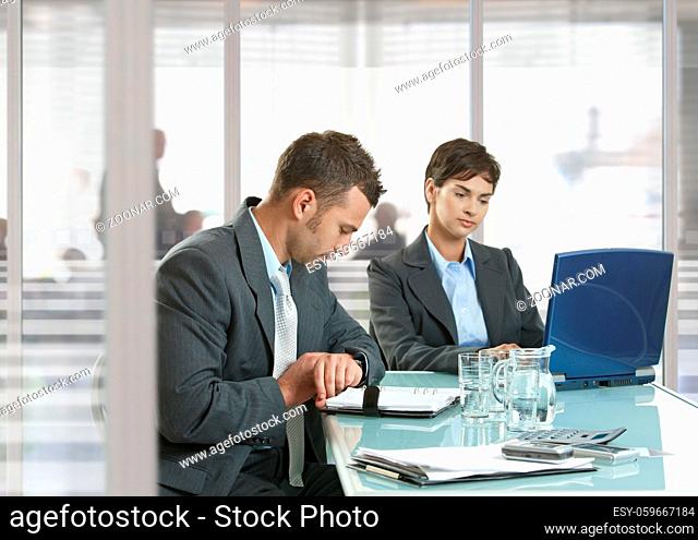 Businessman and businesswoman sitting at desk in office, using laptop computer and checking time