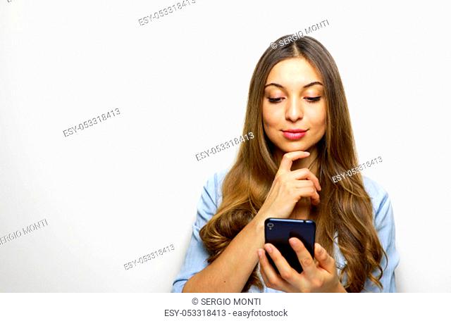 Portrait of a pensive young woman holding and looking her mobile phone with hand on her chin isolated over white background