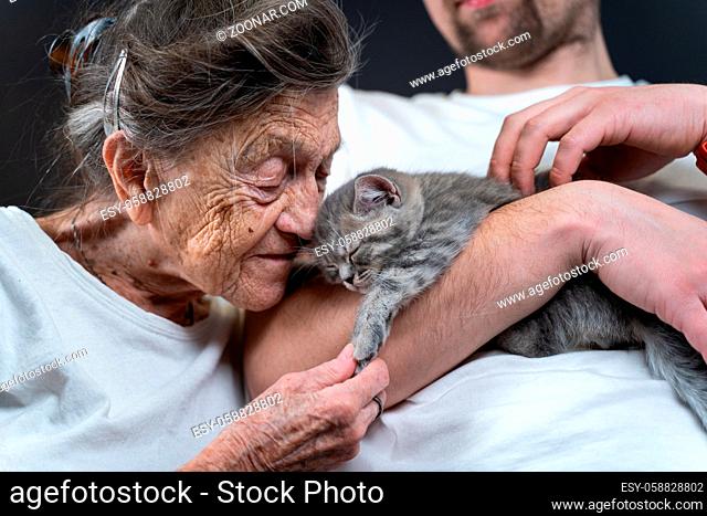 Happy senior woman cuddling and kiss, snuggle up to face small cute gray kitten, which held in arms by his grandson during visit to his grandmother at nursing...