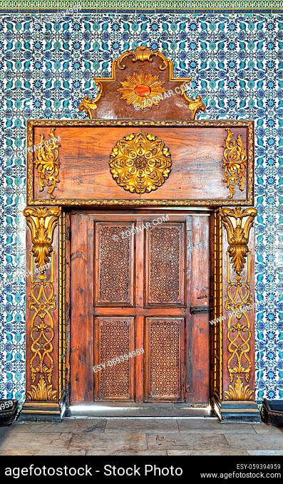 Closed wooden engraved aged door framed by golden ornate wooden frame on Turkish ceramic tiles wall with floral blue patterns