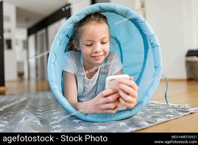 Portrait of smiling girl lying in blue fabric tunnel on the floor at home looking at cell phone