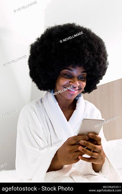 Afro woman in bathrobe using smart phone in bedroom at home