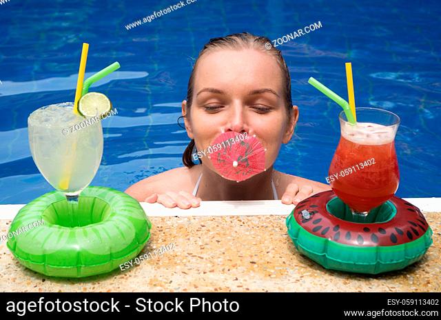Happy smiling woman standing in the water with umbrella in her mouth and cocktails at the edge of the outdoor pool