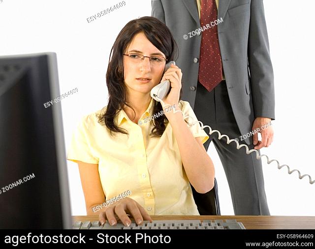 Young businesswoman sitting at desk, talking on phone. Working on computer. Man in suit standing behind her. White background