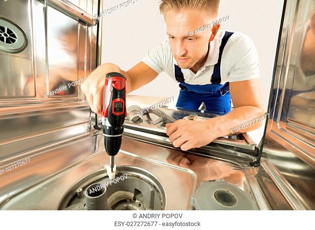 Young Man In Overall Repairing Dishwasher With Electric Drill
