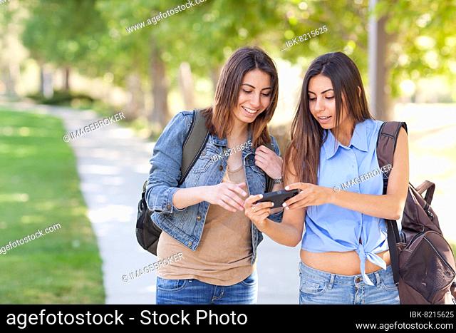 Two beautiful young ethnic twin sisters with backpacks using A smartphone outside