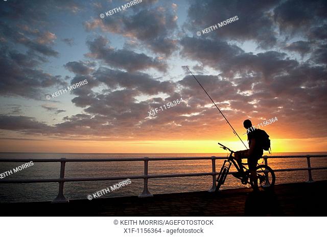 The silhouette of a young man on a bicycle at sunset, about to go fishing, cardigan bay west wales UK