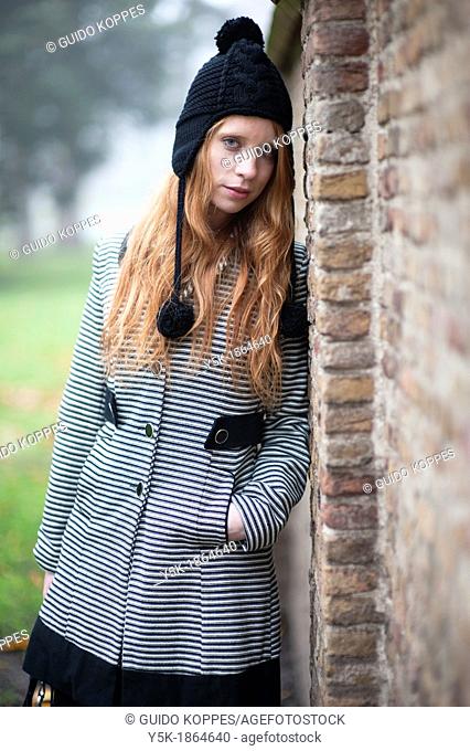 Breda, Netherlands. Young, fashionable and redheaded woman in a city-park, leaning against a wall