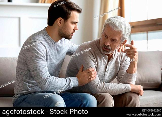 Young stressed man comforting irritated mature old father, sitting together on couch. Worried grownup son feeling sorry, asking forgiveness to angry offended...