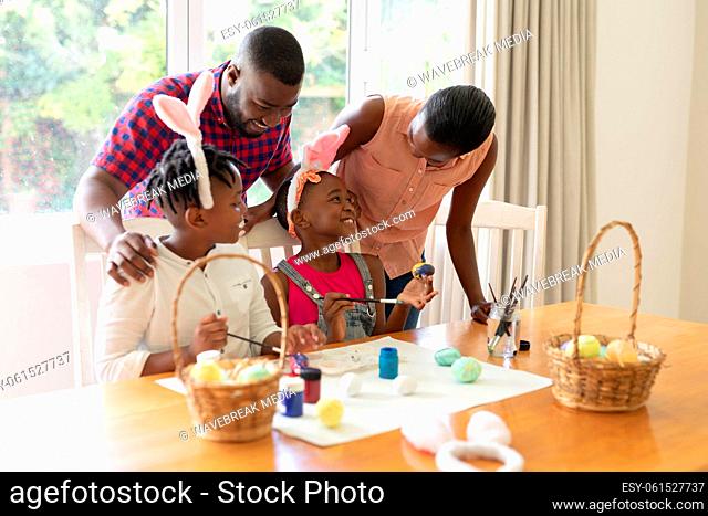 Happy african american parents with son and daughter wearing bunny ears painting colourful eggs