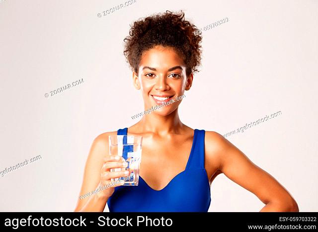 Close-up of young happy african-american fitness woman, holding glass of water and smiling pleased, standing against white background