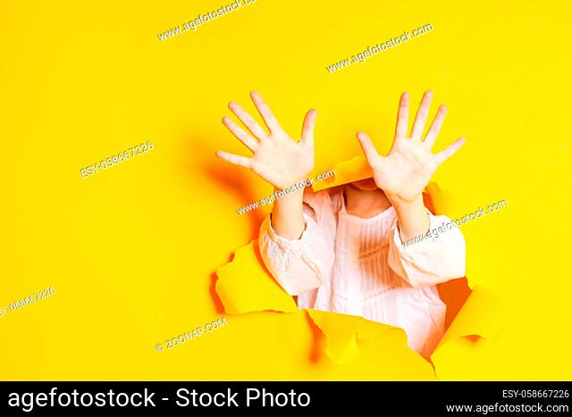 Child hands is showing ten fingers through a ripped hole in yellow paper, with copy space. Gestures and body language