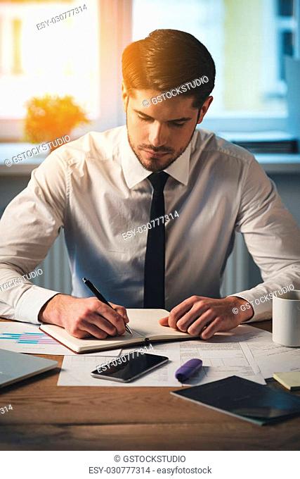 Making some notes. Pensive young handsome man writing in his notebook while sitting at his working place