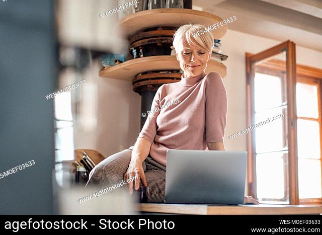 Female freelance worker with laptop sitting on kitchen counter at home