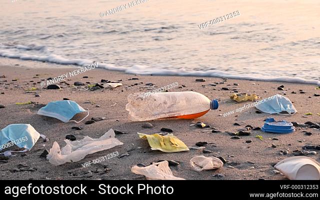 Disposable face masks and plastic debris on the beach in surf zone. Coronavirus COVID-19 is contributing to pollution, as discarded used masks clutter polluting...